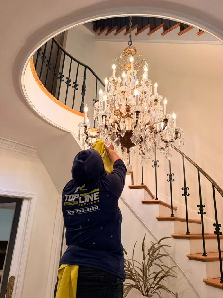 Top of the Line technician hand-cleaning a crystal chandelier beneath a curved staircase