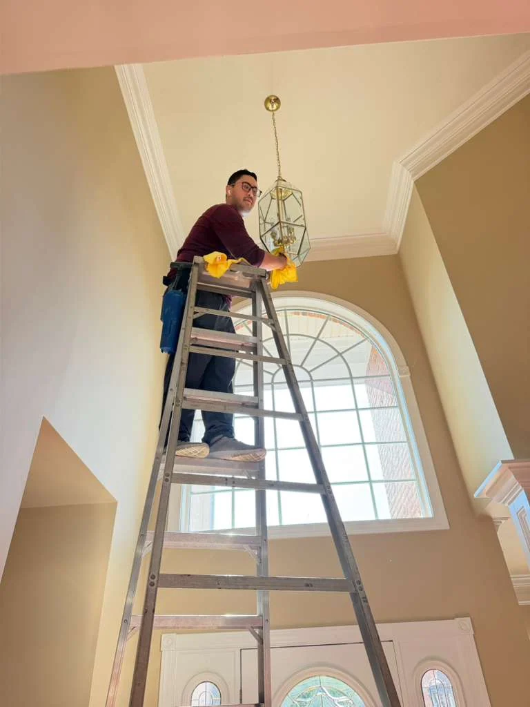 Technician on a ladder cleaning a hanging light fixture in a tall foyer