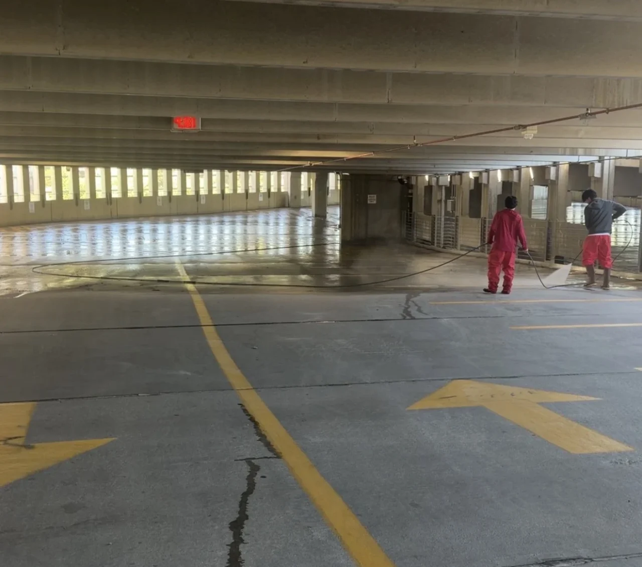 Two technicians pressure washing the floor of a covered parking garage.