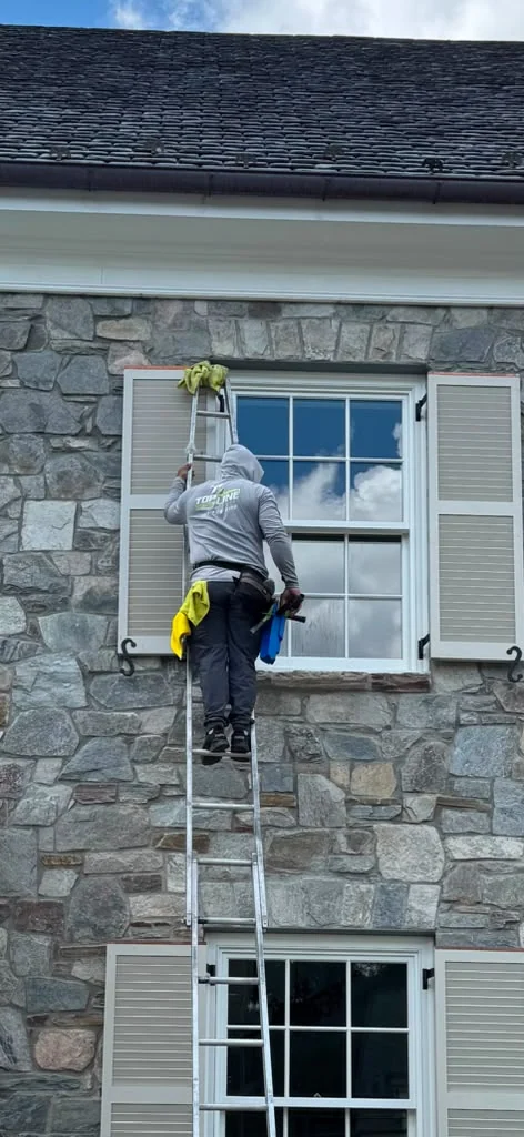 Technician on a ladder cleaning an upper-story window on a stone home